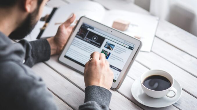 A man uses a tablet to browse news while enjoying a cup of coffee, stylus in hand.
