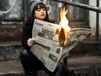 Woman sits outdoors reading a newspaper on fire, creating a dramatic urban scene.