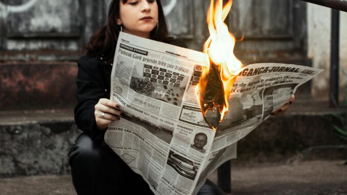 Woman sits outdoors reading a newspaper on fire, creating a dramatic urban scene.