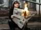 Woman sits outdoors reading a newspaper on fire, creating a dramatic urban scene.