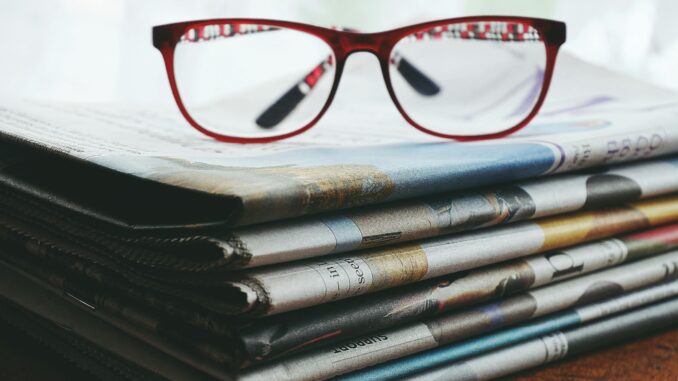 Red eyeglasses resting on a stack of newspapers symbolize reading and information.