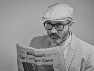 A man in a suit, wearing glasses and a cap, reads The New York Times.