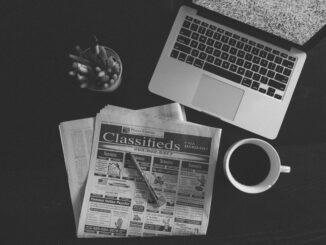 A black and white image of a laptop, newspaper, and coffee cup on a desk.