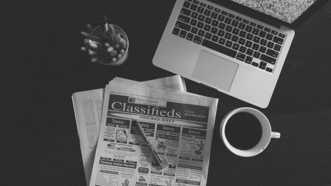 A black and white image of a laptop, newspaper, and coffee cup on a desk.