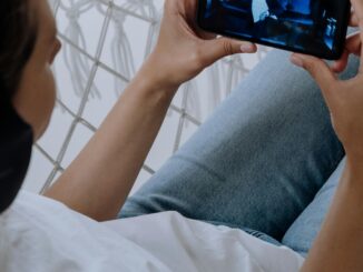 A woman in blue jeans and headphones watches a video on her smartphone while relaxing indoors.