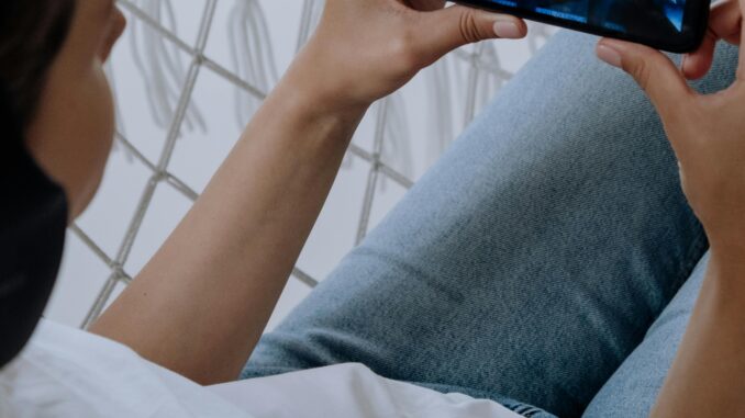 A woman in blue jeans and headphones watches a video on her smartphone while relaxing indoors.