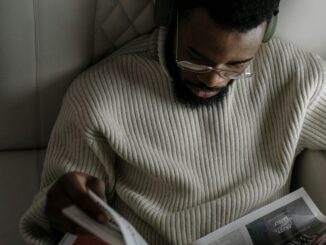 A man with facial hair wearing headphones and glasses reads a newspaper in a cozy setting.