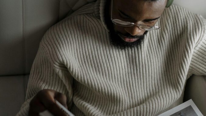 A man with facial hair wearing headphones and glasses reads a newspaper in a cozy setting.