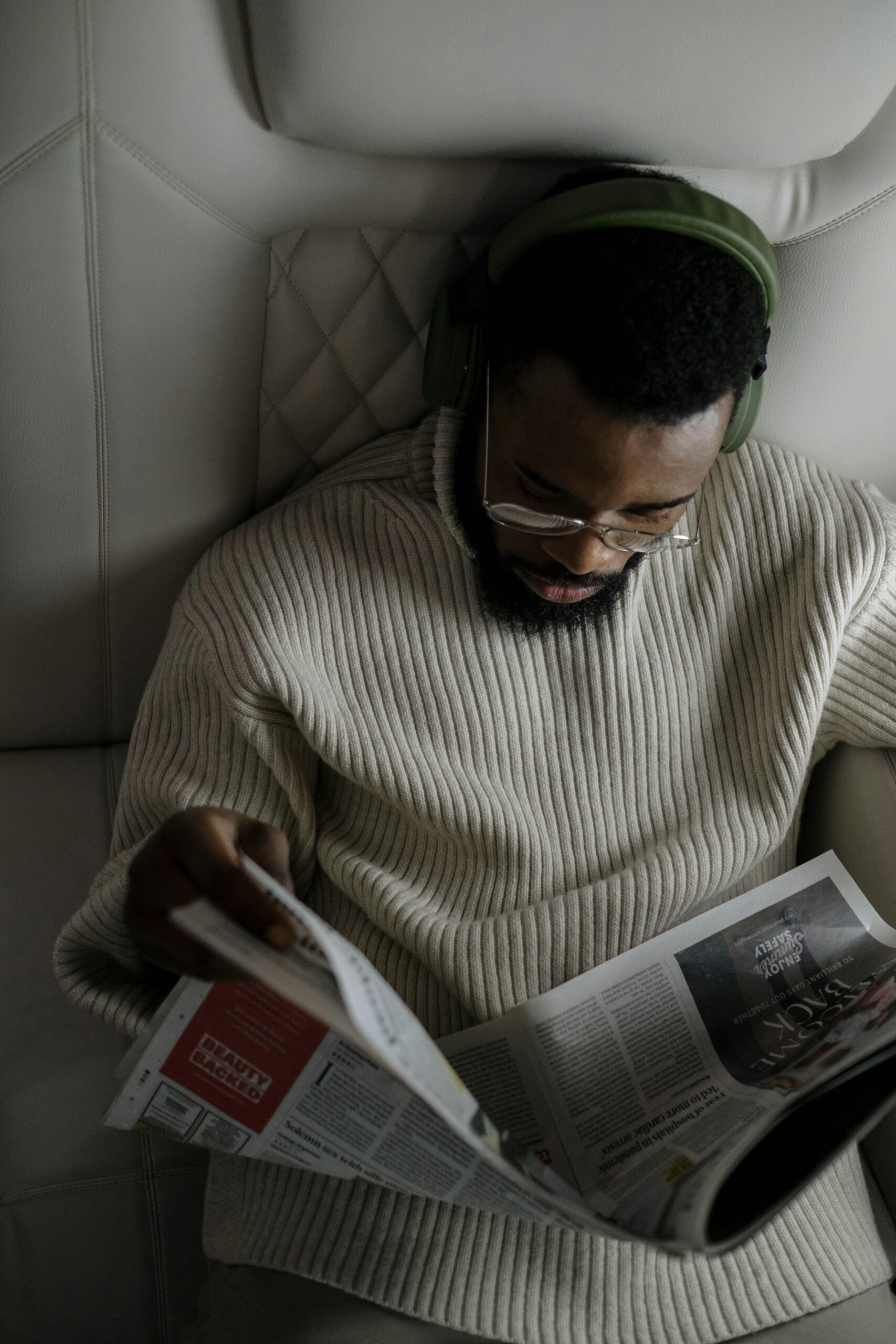 A man with facial hair wearing headphones and glasses reads a newspaper in a cozy setting.