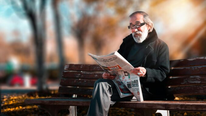 Senior man with beard reading a newspaper on a wooden bench in a sunny park setting.