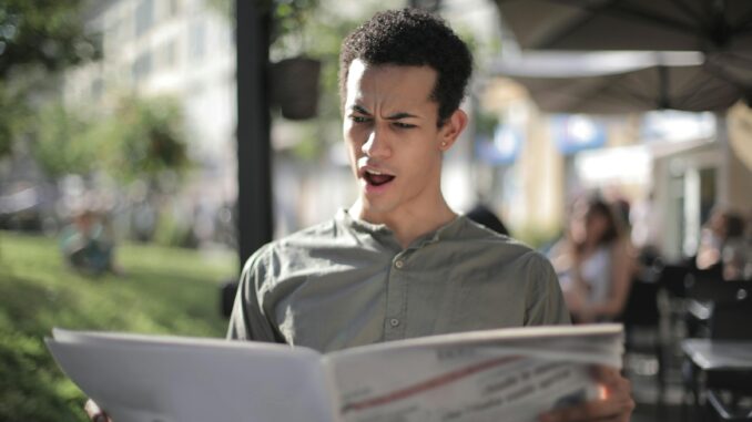 A young man is surprised while reading a newspaper at an outdoor cafe.