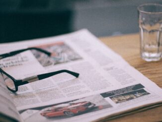 Eyewear on magazine with text in columns and illustrations near cup with saucer on wooden table in cafeteria