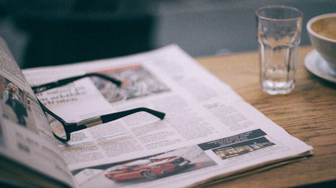 Eyewear on magazine with text in columns and illustrations near cup with saucer on wooden table in cafeteria