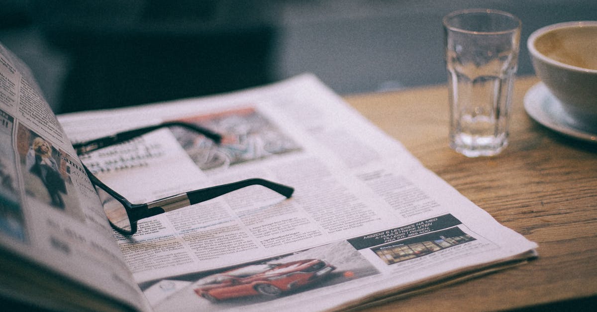 Eyewear on magazine with text in columns and illustrations near cup with saucer on wooden table in cafeteria