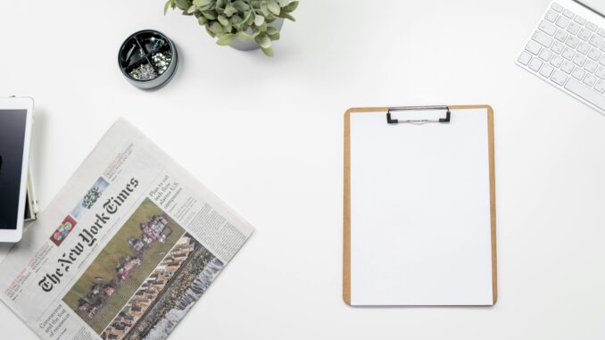 Clean flatlay of a home office desk with The New York Times, clipboard, and keyboard.