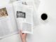 A close-up shot of a hand holding a newspaper with a coffee cup on a white surface.