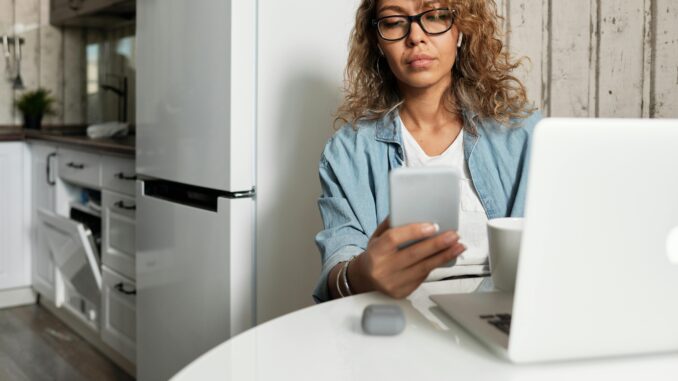 Woman using smartphone and laptop while working remotely in a home kitchen setting.