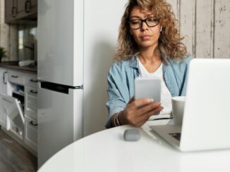 Woman using smartphone and laptop while working remotely in a home kitchen setting.