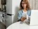 Woman using smartphone and laptop while working remotely in a home kitchen setting.