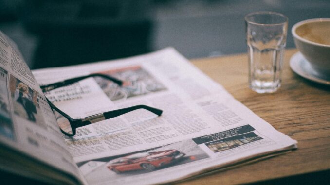Eyewear on magazine with text in columns and illustrations near cup with saucer on wooden table in cafeteria