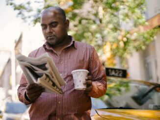 Interested adult ethnic male taxi driver with disposable glass of hot drink reading newspaper near vehicle on city street