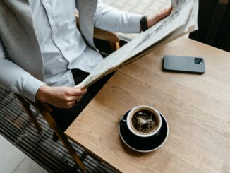 An adult male enjoying his coffee while reading a newspaper at a cafe with his smartphone nearby.