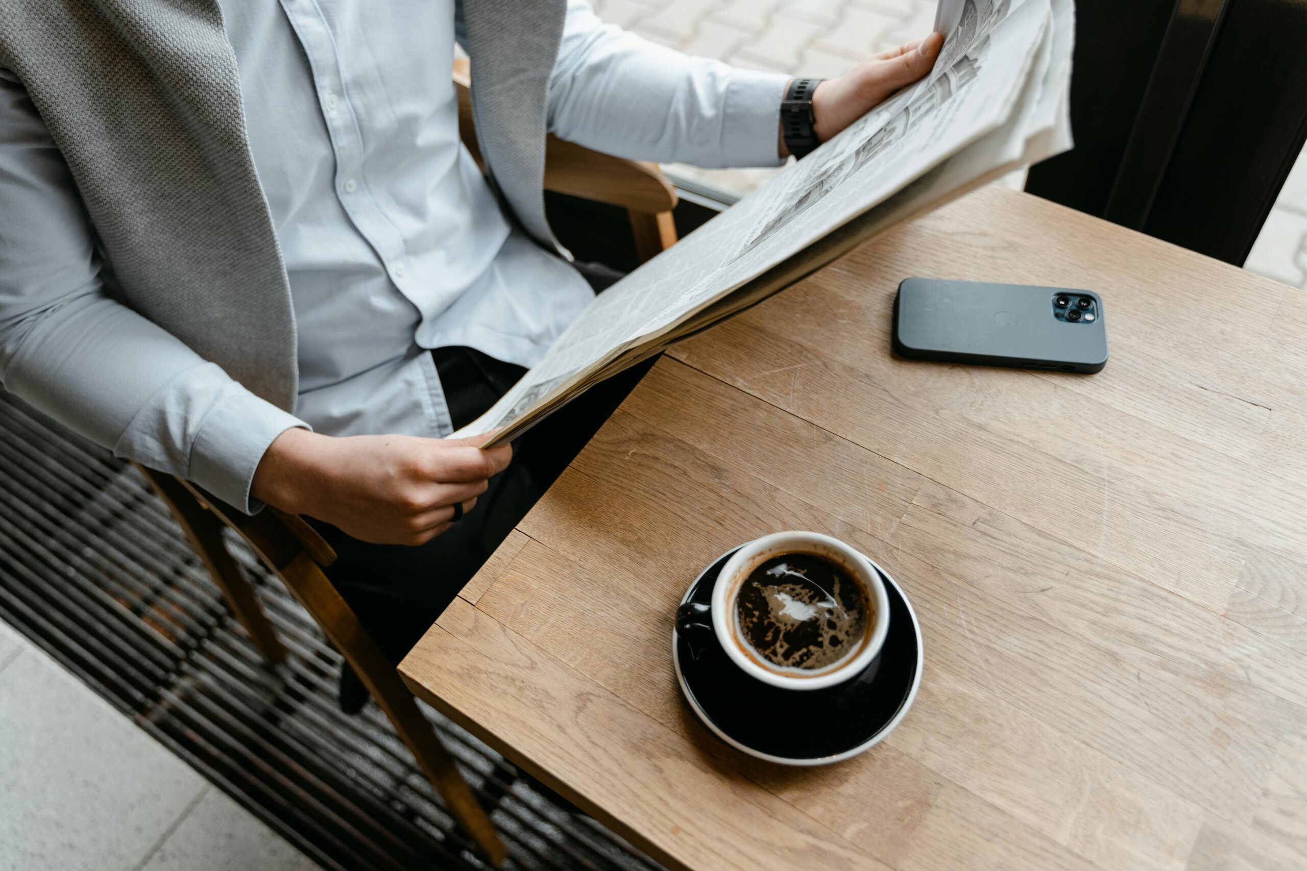 An adult male enjoying his coffee while reading a newspaper at a cafe with his smartphone nearby.