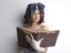 A woman wearing a vintage hat and gloves reads an old book indoors.