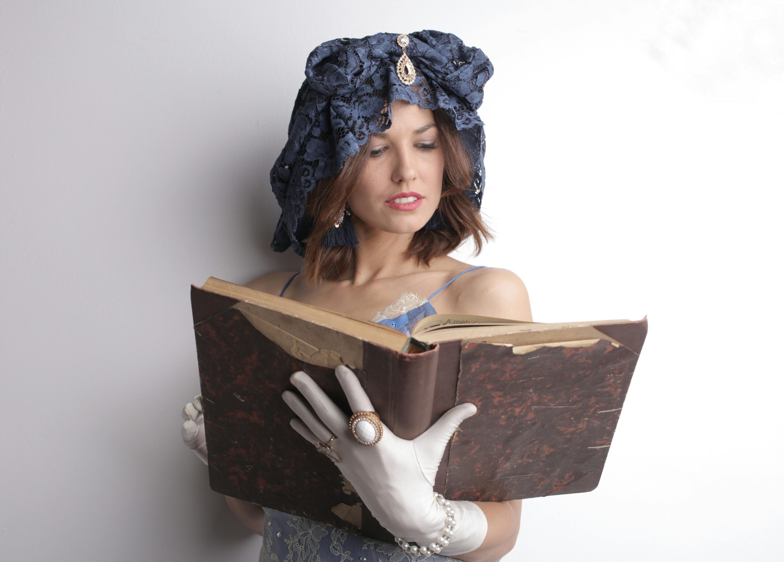 A woman wearing a vintage hat and gloves reads an old book indoors.