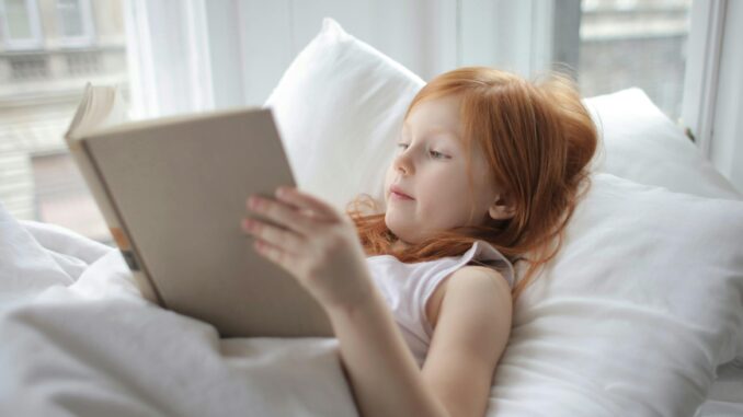 A young girl with red hair reading a book comfortably in bed, enjoying leisure time indoors.