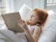 A young girl with red hair reading a book comfortably in bed, enjoying leisure time indoors.