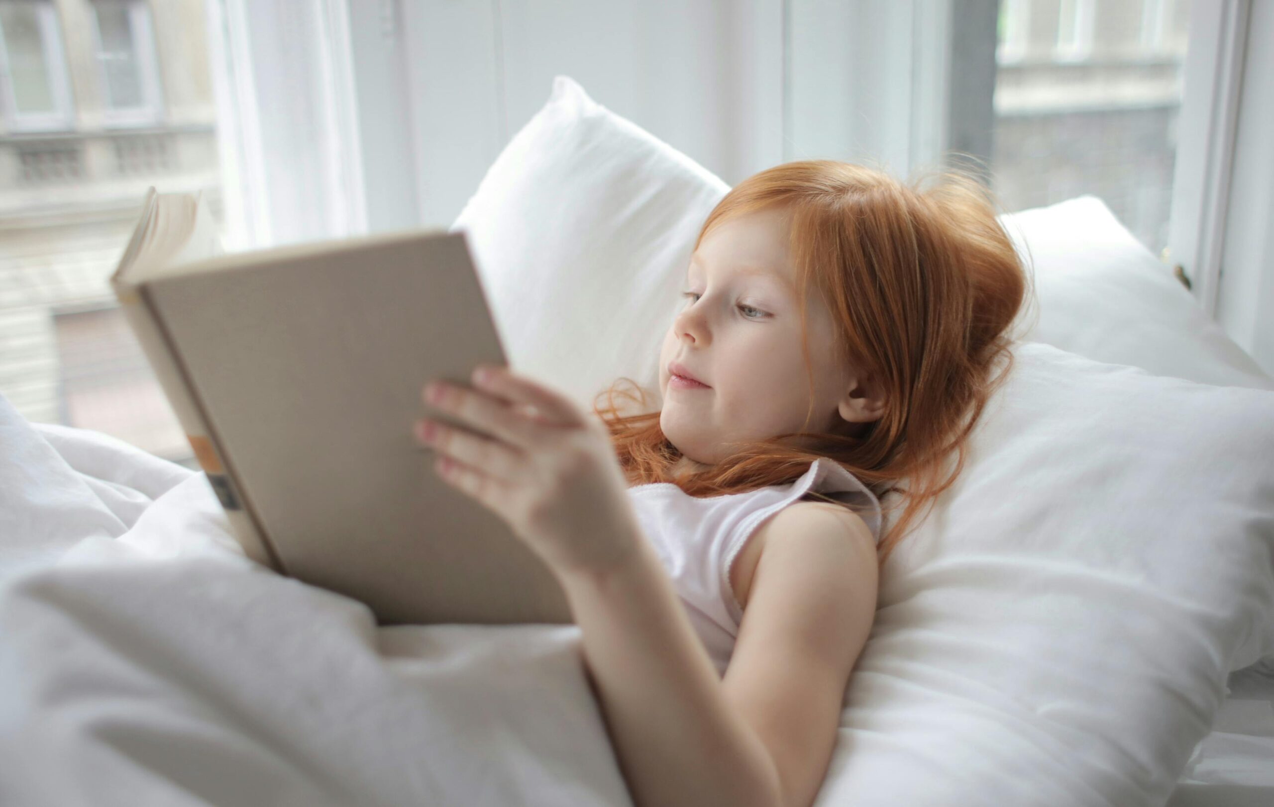 A young girl with red hair reading a book comfortably in bed, enjoying leisure time indoors.