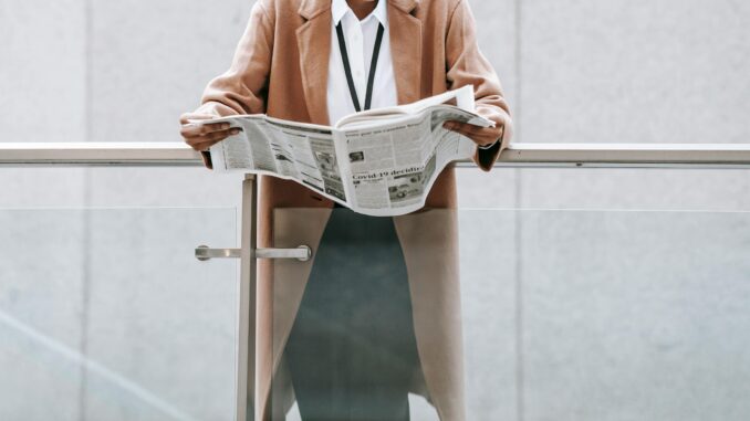 Crop content African American businesswoman in formal clothes reading fresh newspaper and leaning on glass railing of contemporary office building
