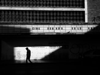 Black and white photo of a man walking in front of an old urban theater, creating dramatic shadows.