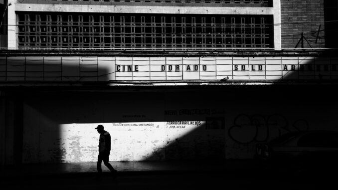 Black and white photo of a man walking in front of an old urban theater, creating dramatic shadows.