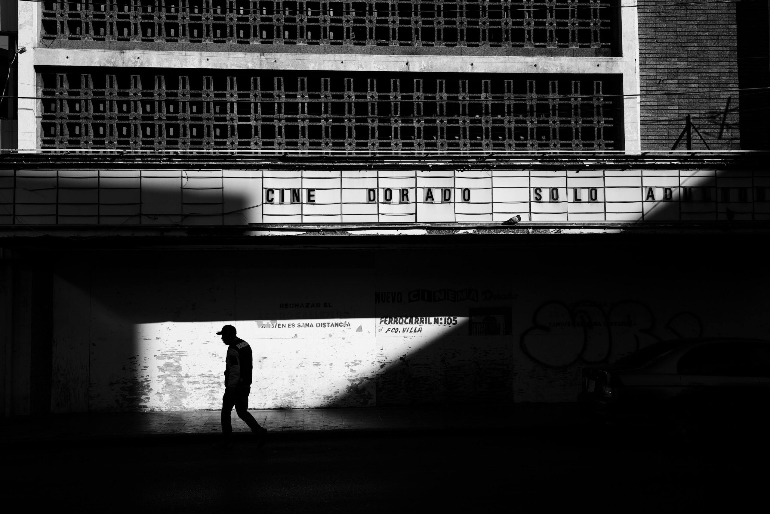 Black and white photo of a man walking in front of an old urban theater, creating dramatic shadows.