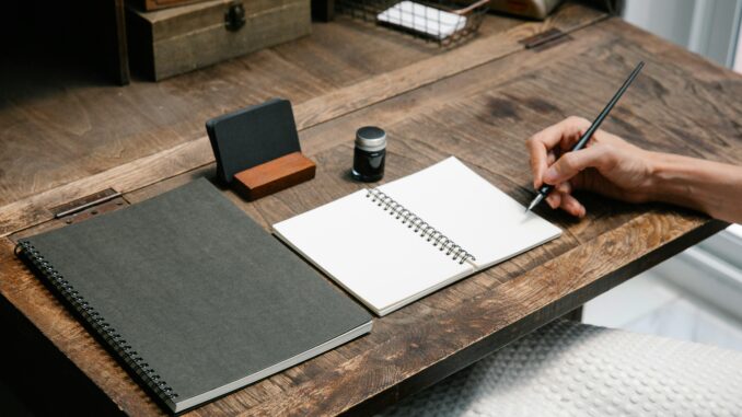 A close-up of a hand writing in a notebook at a tidy wooden desk, evoking creativity.