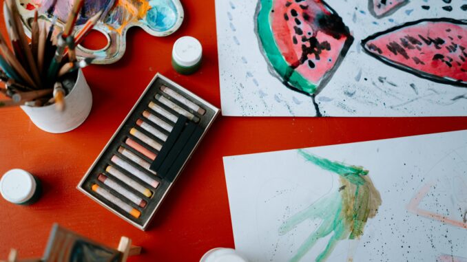 Creative art setup featuring watercolors and pastels on a red table, showcasing artistic expression.