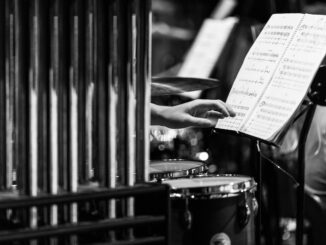 Monochrome image of a hand reaching for sheet music in a percussion setup.