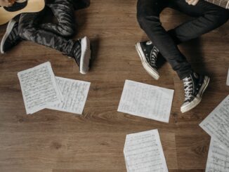 Two musicians playing guitars surrounded by sheet music on a wooden floor.