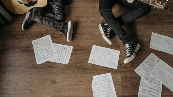 Two musicians playing guitars surrounded by sheet music on a wooden floor.