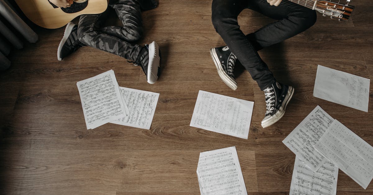 Two musicians playing guitars surrounded by sheet music on a wooden floor.