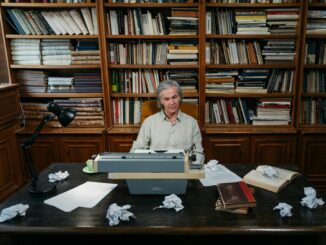 Senior man at a typewriter in a library, surrounded by books and paper.