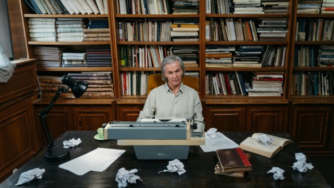 Senior man at a typewriter in a library, surrounded by books and paper.