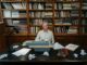 Senior man at a typewriter in a library, surrounded by books and paper.