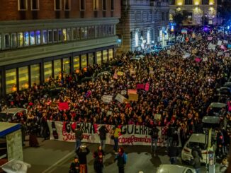 A massive nighttime protest with crowds holding banners and placards on a city street.