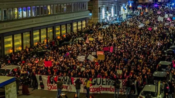 A massive nighttime protest with crowds holding banners and placards on a city street.