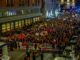 A massive nighttime protest with crowds holding banners and placards on a city street.