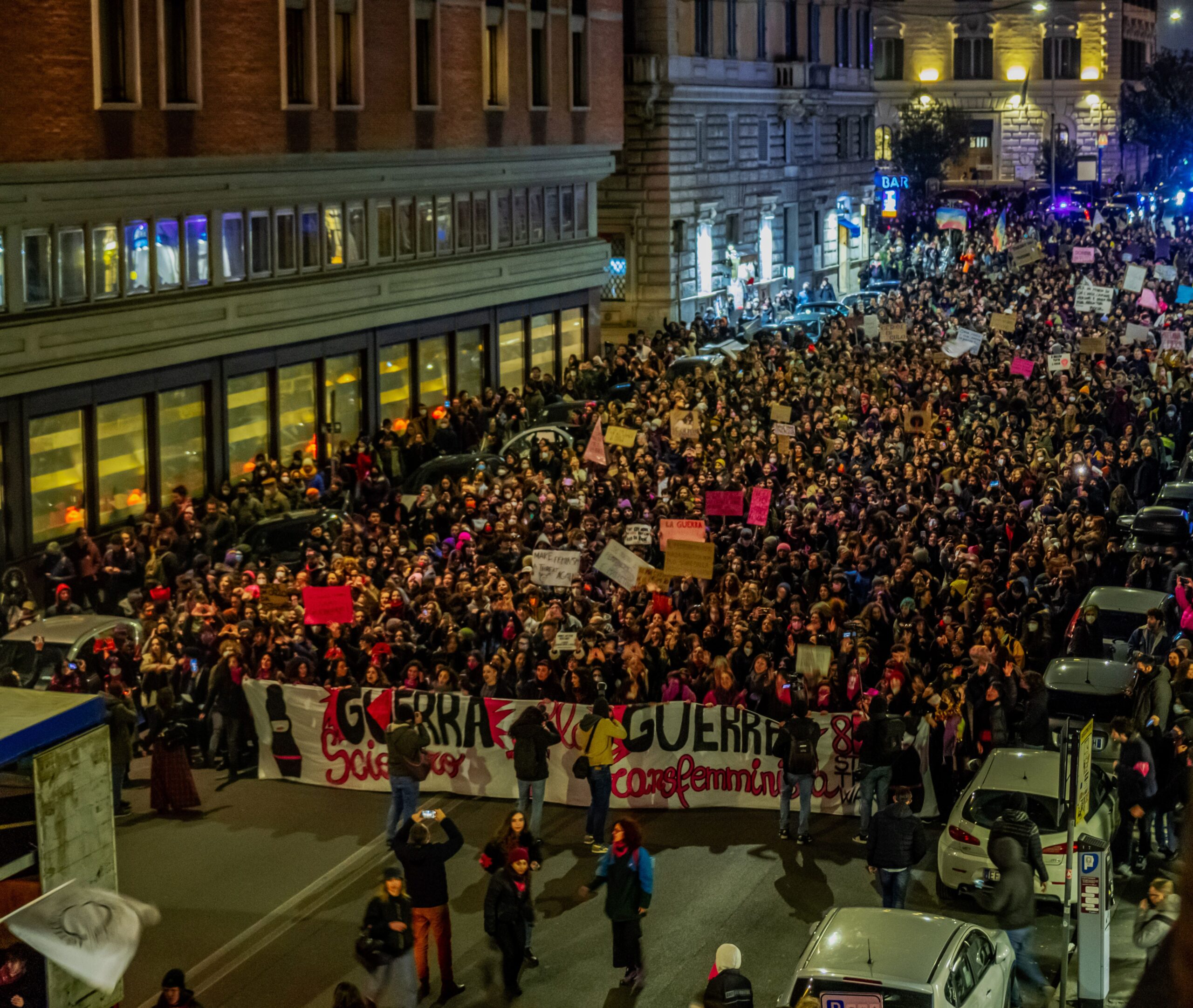 A massive nighttime protest with crowds holding banners and placards on a city street.
