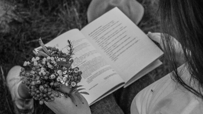 A person reads a book while holding flowers, captured in black and white outdoors.